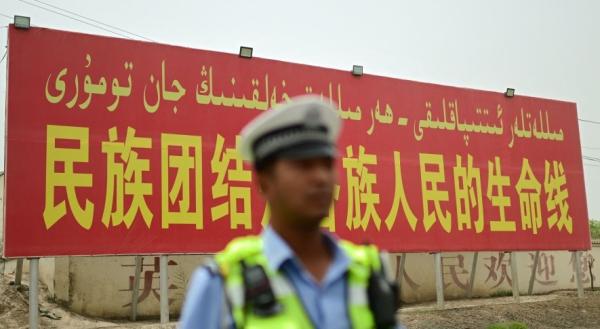 2023年7月19日的照片显示,在中国西北部新疆地区喀什地区的一条道路上,一名警察站在一个宣传广告牌前,上面写着 “ 民族团结是各族人民的生命线 ” 。(图片来源:翻拍)